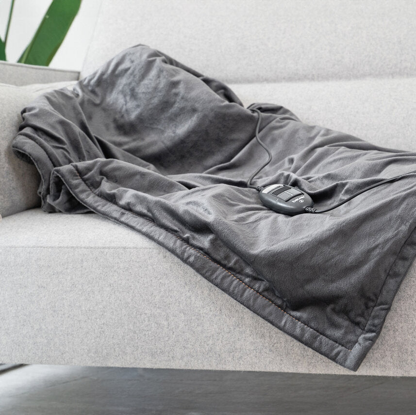 Ein grauer, kuscheliger Schlafsack liegt auf einer Couch, mit einem Kabel und einem kleinen Gerät darauf.