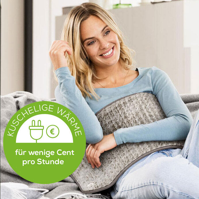 Frau sitzt auf Couch, hält Kissen, lächelt, trägt blauen Pullover, grünes Label mit Text und Symbolen.