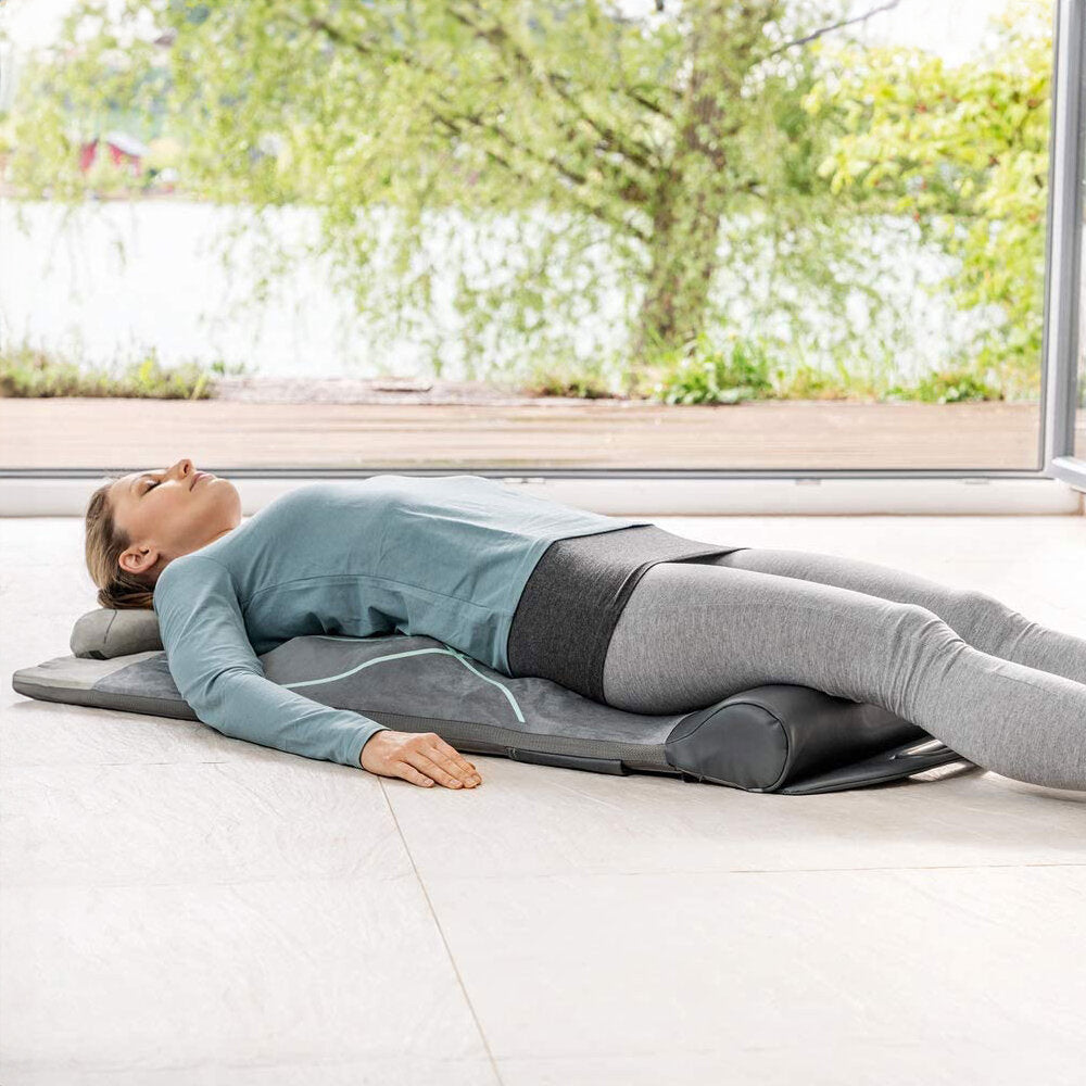 Frau liegt entspannt auf einer Liege mit Kopfstütze vor einem großen Fenster, Blick nach oben, im Innenraum.