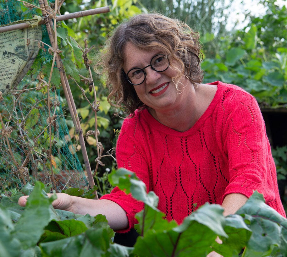 Frau mit Brille und roten Pullover im Garten, umgeben von grünen Pflanzen und Gartenstrukturen.