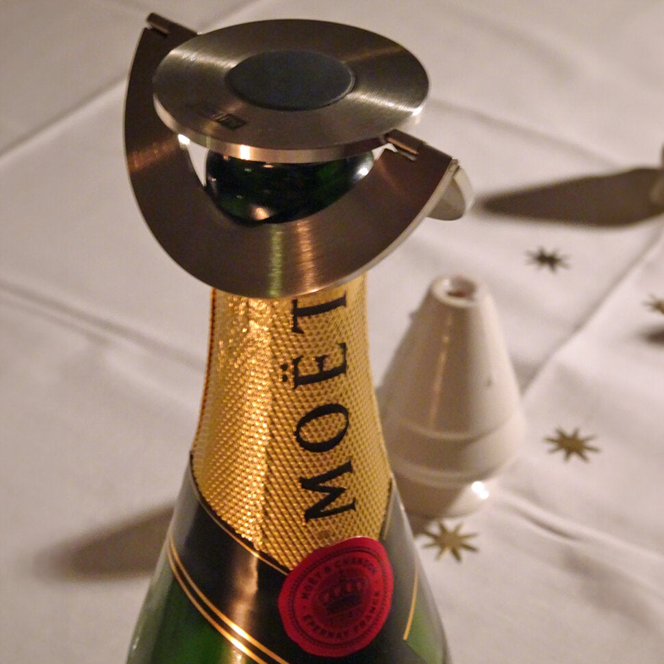 Sektflasche mit Metalldeckel und goldener Banderole, daneben ein weißer Partyhut auf weißem Tisch mit goldenen Sternen.