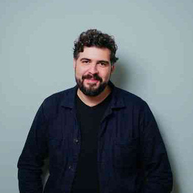 Porträt eines Mannes mit dunklem Haar, Bart und lockigem Haar, trägt dunkle Jacke vor grauem Hintergrund.