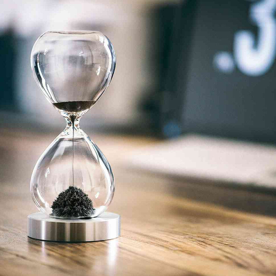 Sanduhr mit schwarzem Sand, auf Holzplatte, im Hintergrund Bildschirm.
