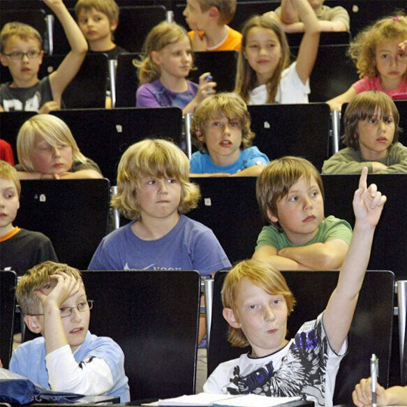 Kinder sitzen in einem Klassenzimmer, einige heben die Hand, andere schauen nach vorne.