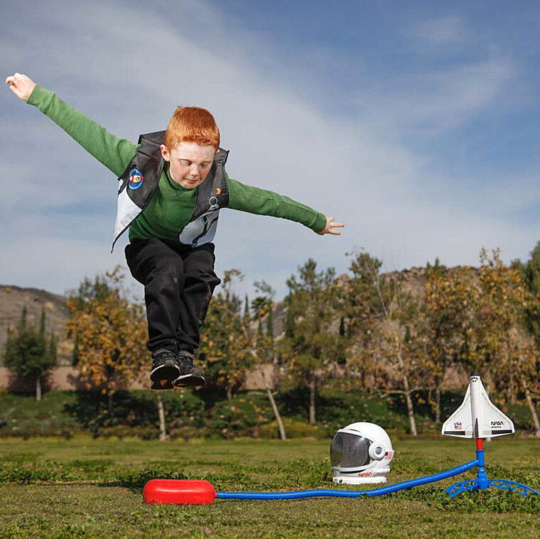 Junge mit Rucksack springt in die Luft vor einem Raketen-Spielzeug auf dem Rasen.