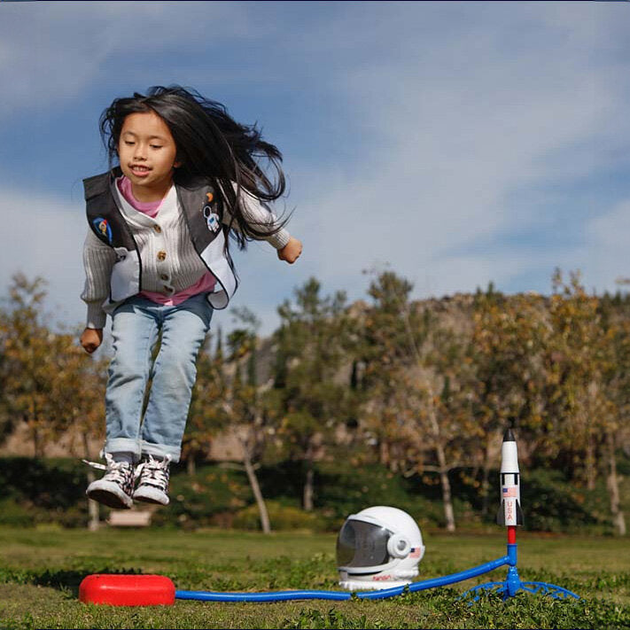Mädchen springt in einem Park, mit Raumfahrt-Helm und Raketenmodell im Hintergrund.