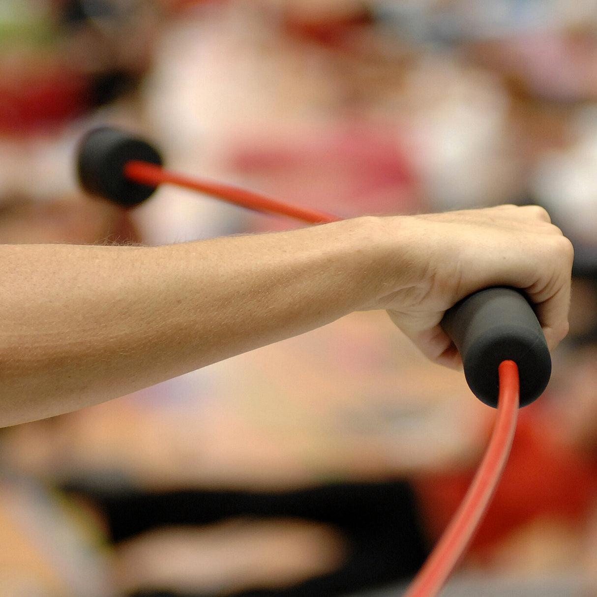 Arm hält einen roten Widerstand mit schwarzen Endkappen, im Hintergrund unscharfe Personen.