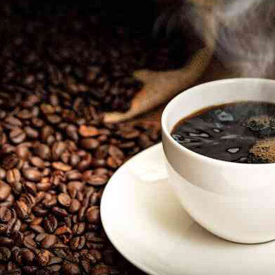 Eine weiße Kaffeetasse mit schwarzem Kaffee auf einem weißen Unterteller, daneben Kaffeebohnen und eine dampfende Kaffeetasse.