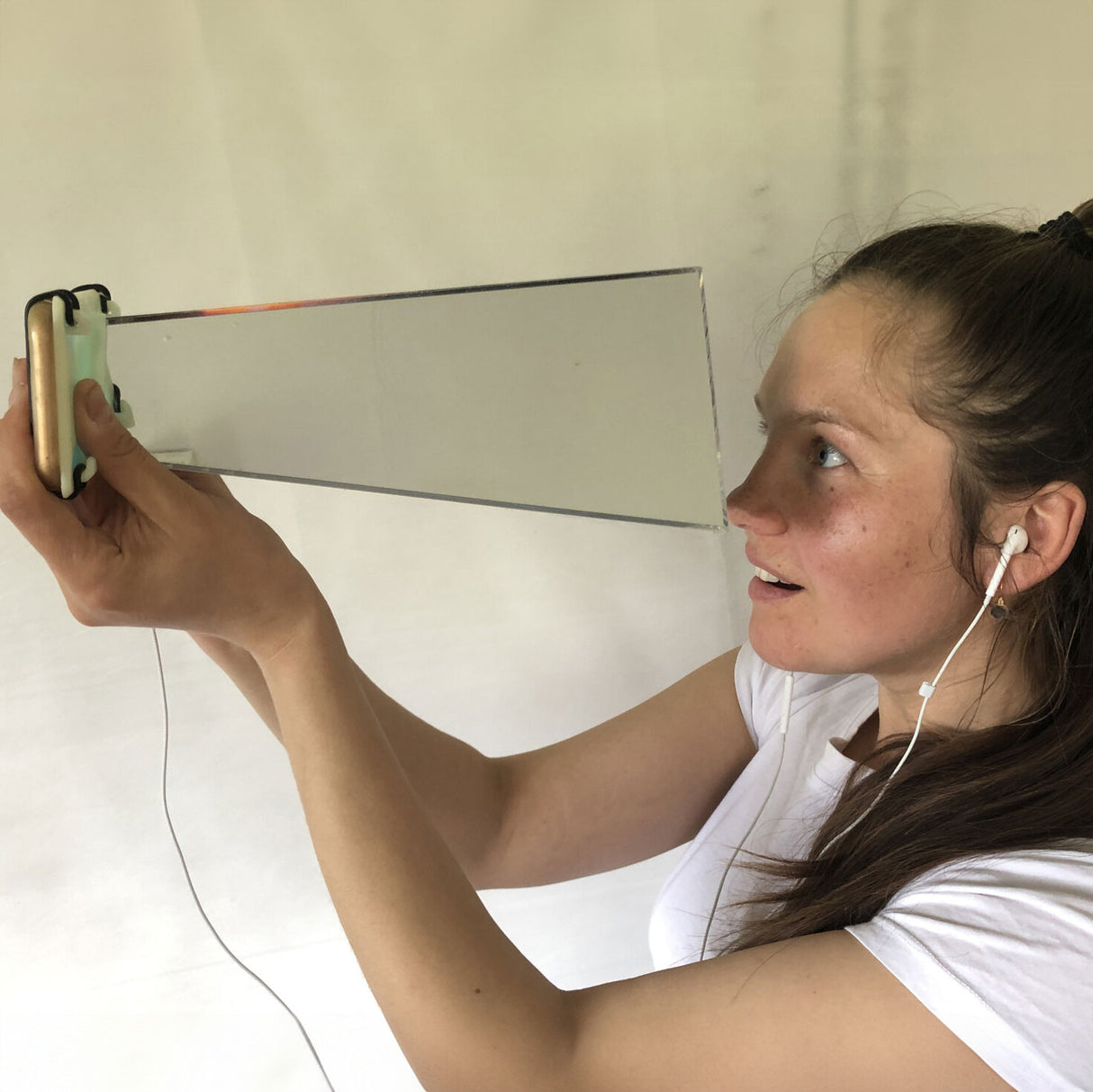 Frau hält einen Spiegel, trägt Ohrhörer, schaut nach oben, hat braune Haare, trägt ein weißes T-Shirt.
