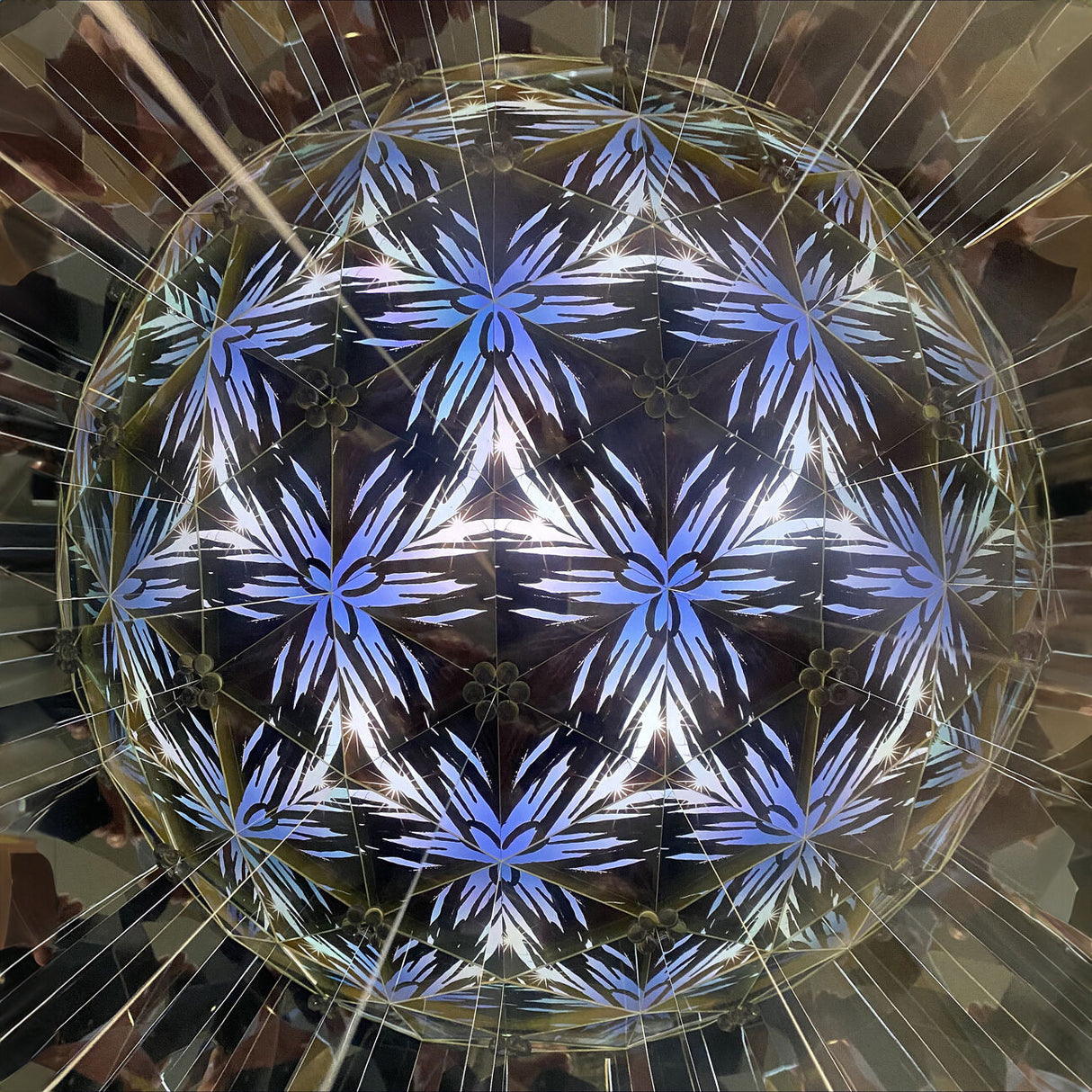Blick nach oben auf eine geometrische, leuchtende Kugel mit blauen und weißen Mustern, umgeben von reflektierenden Flächen.