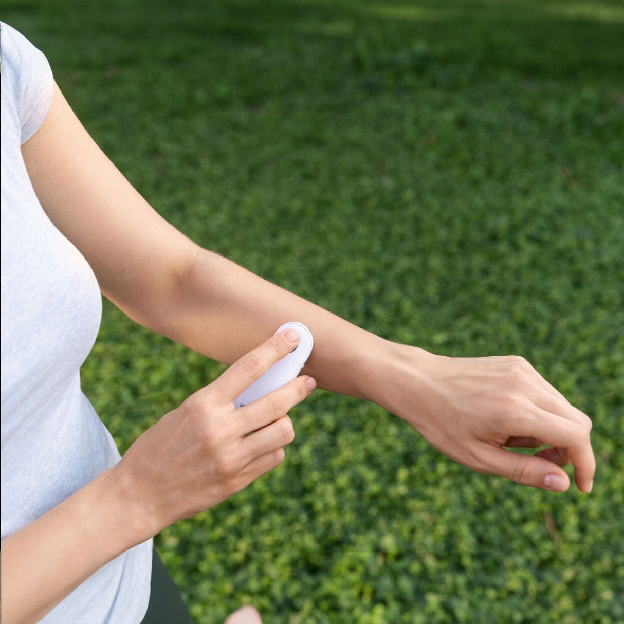 Person benutzt ein Handgerät zur Hautpflege an ihrem Arm, im Freien auf grünem Rasen.