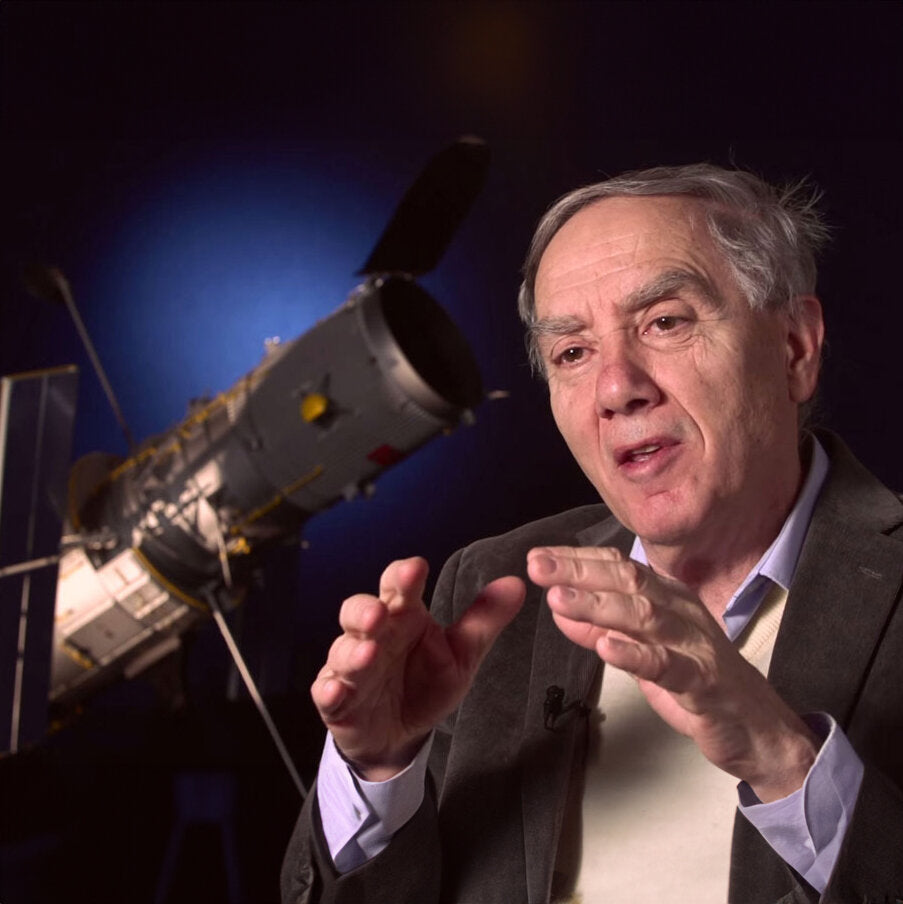 Älterer Mann erklärt mit Handgesten vor einem Satelliten im Hintergrund.