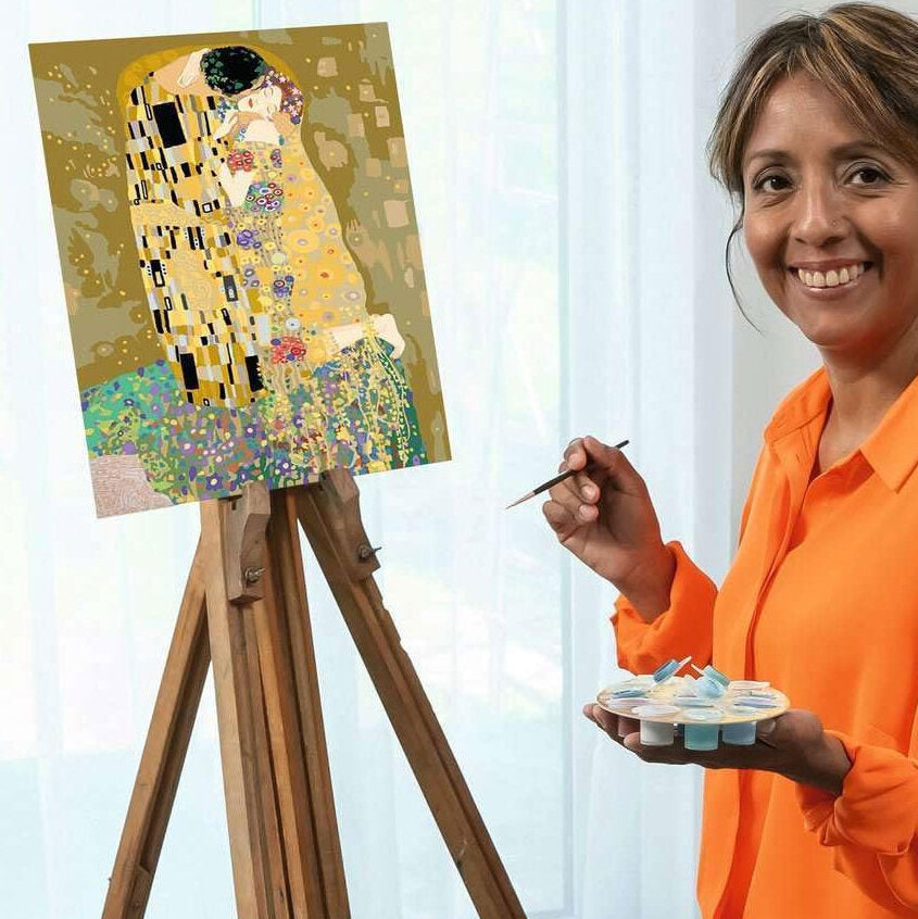 Frau malt ein Bild im Stil von Gustav Klimt auf einer Staffelei, trägt orange Hemd, hält Pinsel und Farbpalette.