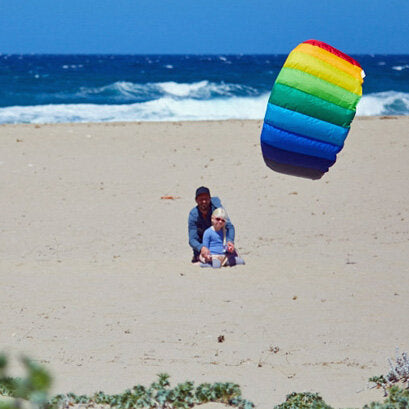 Ein Kind sitzt mit einem Erwachsenen am Strand, im Hintergrund das Meer, und ein farbenfroher, aufgespannter Kite.