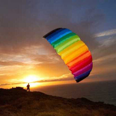 Person mit einem bunten Fallschirm steht auf einem Hügel bei Sonnenuntergang am Meer.