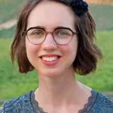 Ein Porträt einer jungen Frau mit Brille, braunen Haaren und einem blauen Oberteil vor grünem Hintergrund.