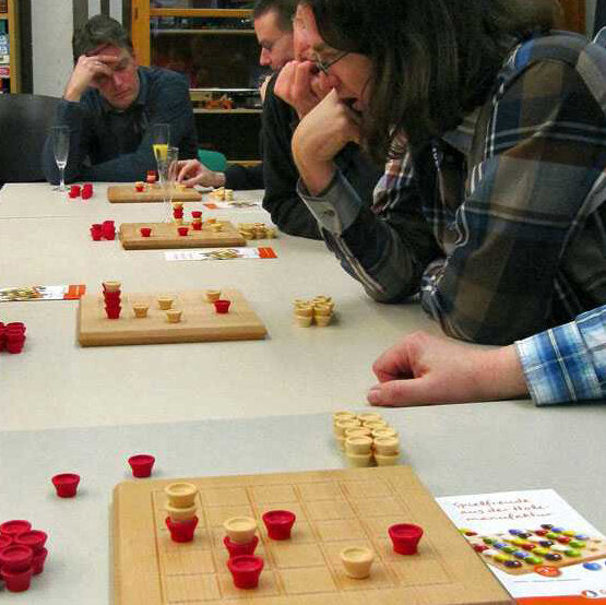 Vier Personen spielen ein Brettspiel mit roten und weißen Spielsteinen an einem Tisch.
