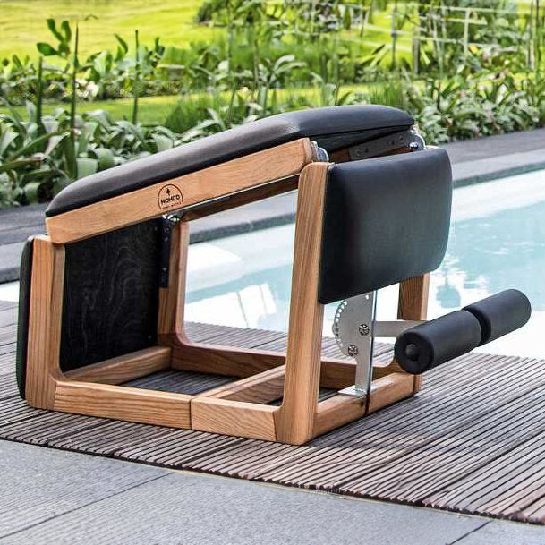 Fitnessgerät aus Holz und Polster neben Pool auf Holzdeck, mit grüner Gartenlandschaft im Hintergrund.