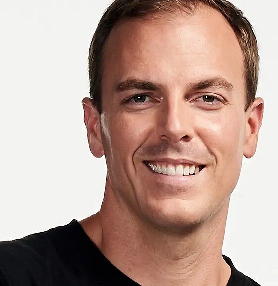 Ein Mann mit kurzem braunem Haar, lächelnd, trägt ein schwarzes Shirt, vor hellem Hintergrund.