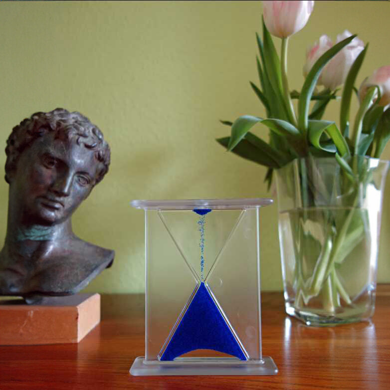 Bronzestatue eines Kopfes, Glasvase mit Tulpen, Sandpendel mit blauer Flüssigkeit auf Holztisch vor grünem Hintergrund.