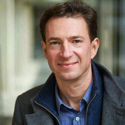 Porträt eines Mannes mit dunklem Haar, blauer Hemd und dunkler Jacke vor einem Gebäude mit Glasfenstern.