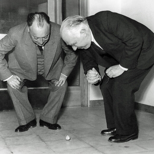 Zwei Männer bücken sich über eine kleine Kugel auf dem Boden, scheinen zu diskutieren oder zu beobachten.