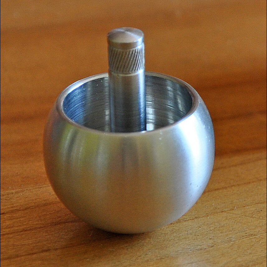 Rundes, silbernes Metallgefäß mit einem vertikalen, geriffelten Metallstab in der Mitte auf einem Holztisch.