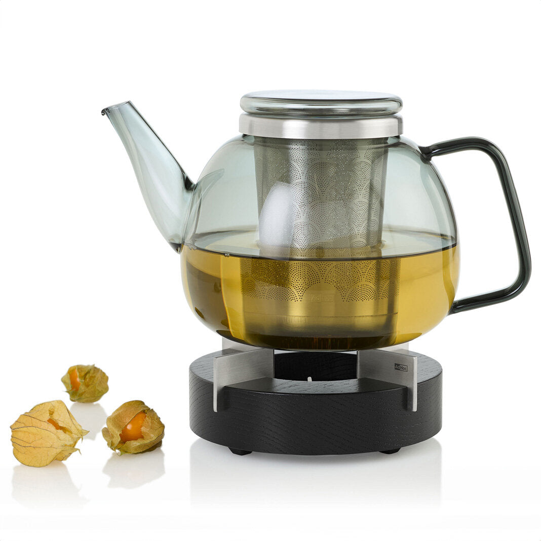 Glas-Teekanne mit gelbem Tee, Teesieb im Inneren, auf einem schwarzen Heizuntersetzer, daneben drei physalis-Früchte.