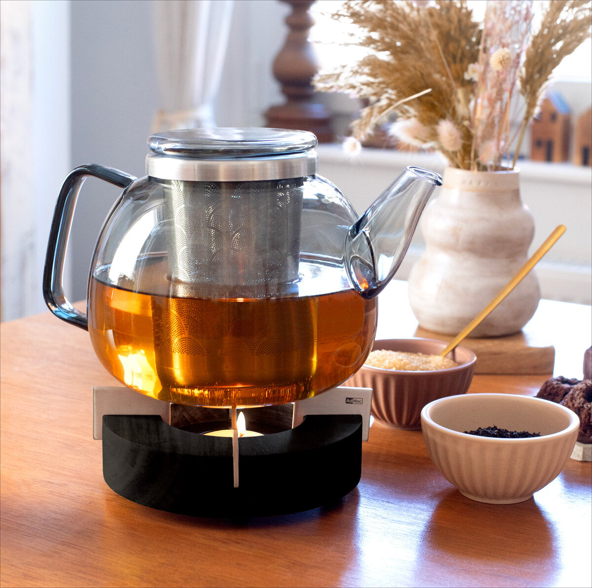 Glas-Kessel mit Tee auf einem Herd, umgeben von Schalen mit Zucker, Keksen und einer Vase mit Trockenblumen.