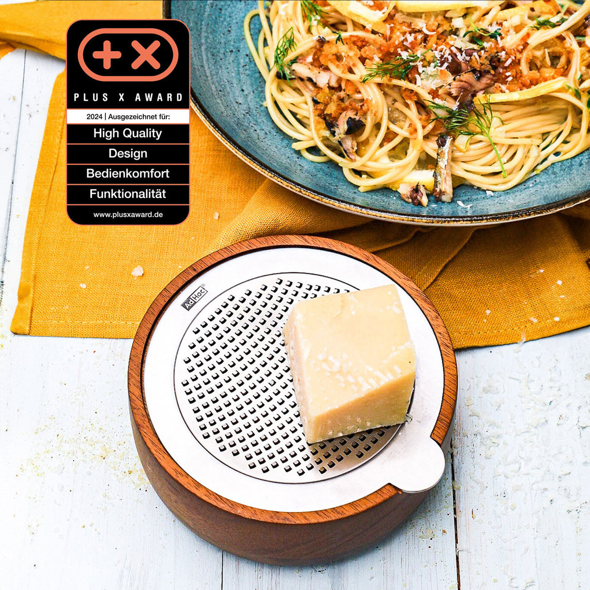 Käsestück auf einer runden Käse-Reibe, daneben Pasta mit Kräutern auf einem blauen Teller, auf gelbem Stoff. Plus X Award-Logo.