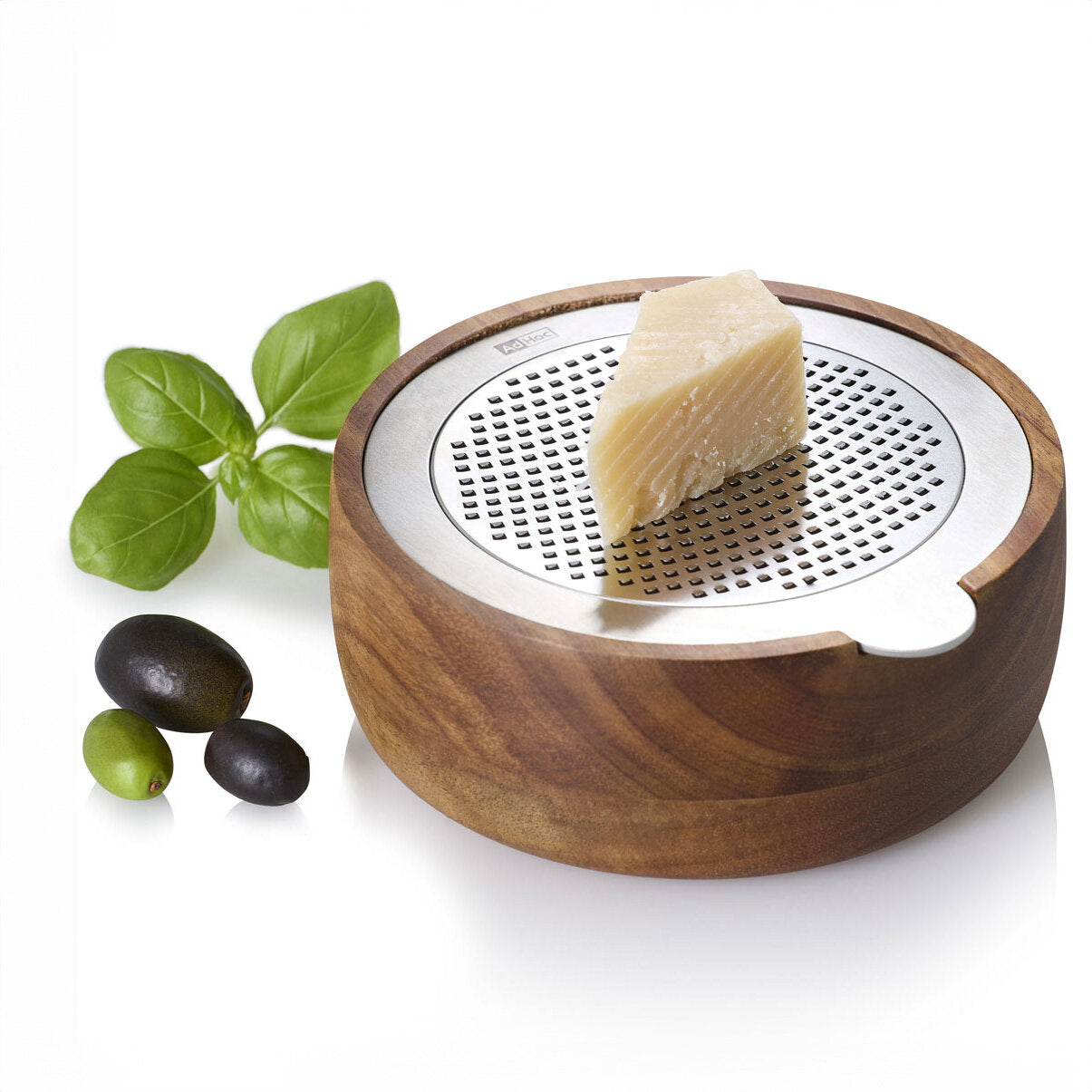 Hartkäse auf Holz- und Metallreibe, daneben Basilikumblätter und schwarze und grüne Oliven.