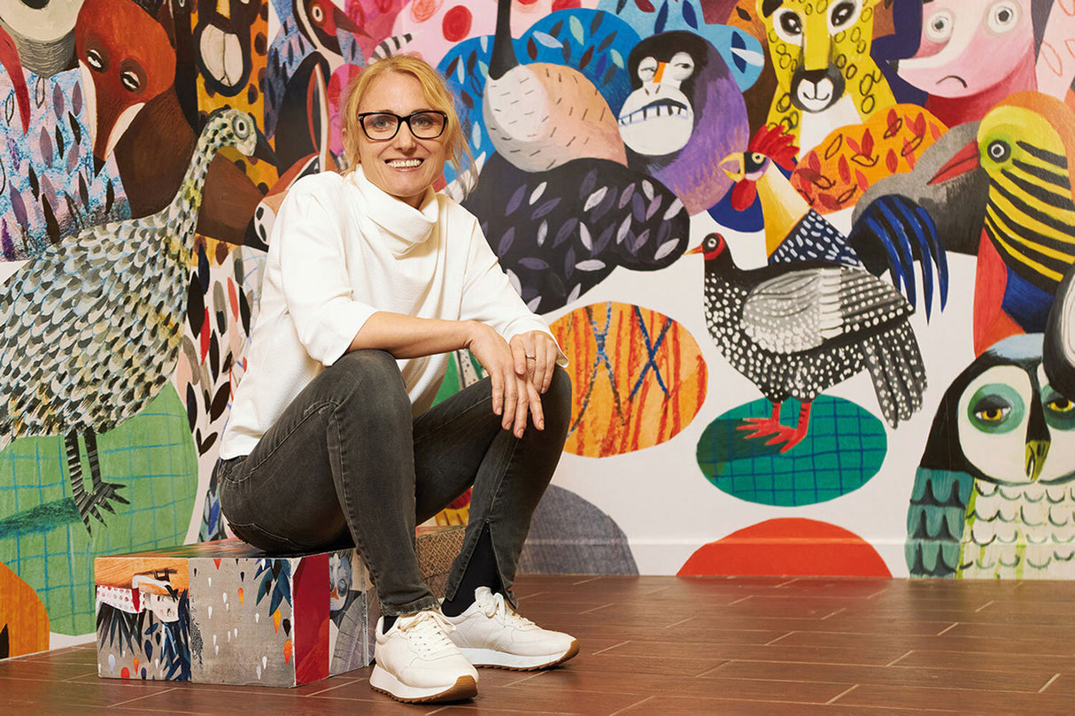 Frau mit Brille sitzt auf bunter, gemalter Bank vor einer Wand mit buntem Tiermural.