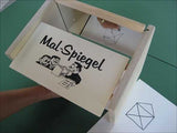 Pappkarton mit "Mal Spiegel" und Cartoon-Illustration, darunter eine Hand, die ein Blatt Papier hält.
