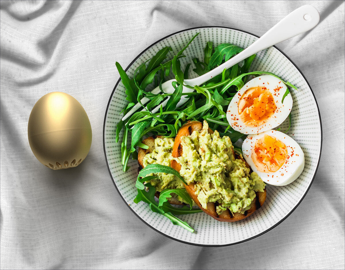 Schüssel mit Salat, Avocado, zwei halbierte Eier mit Paprikapulver, Löffel, goldene Eierform auf weißem Stoff.