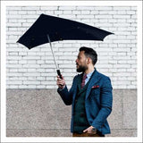 Mann mit dunklem Regenschirm, trägt blaue Jacke, Hemd und Krawatte, steht vor weißer Ziegelwand.