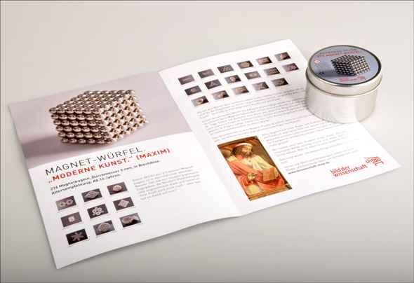 Offener Flyer mit Magnetwürfeln und Text, daneben eine runde Dose mit Magnetwürfeln.