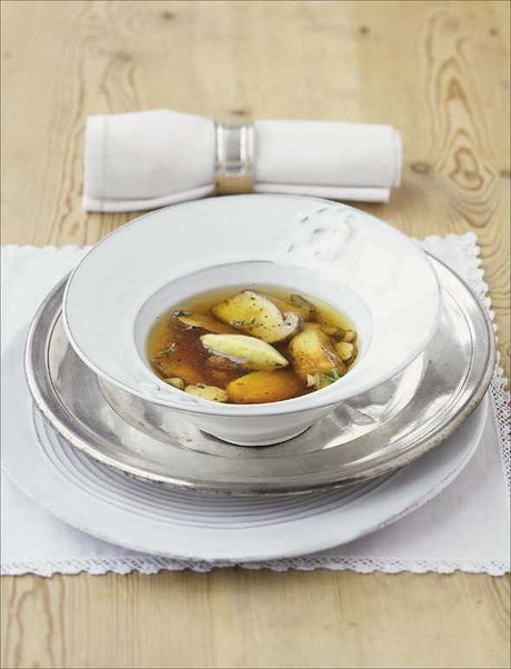 Schüssel mit Suppe, darin Knoblauchzehen und Gewürze, auf einem silbernen Teller, mit Besteckrolle im Hintergrund.