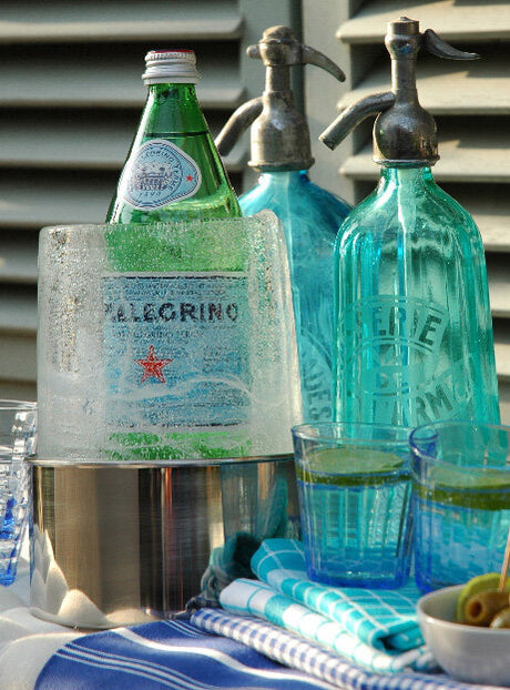 Bunte Wasserflaschen, Gläser mit Getränken, Eis und Servietten auf einem Tisch vor einer Lüftung.