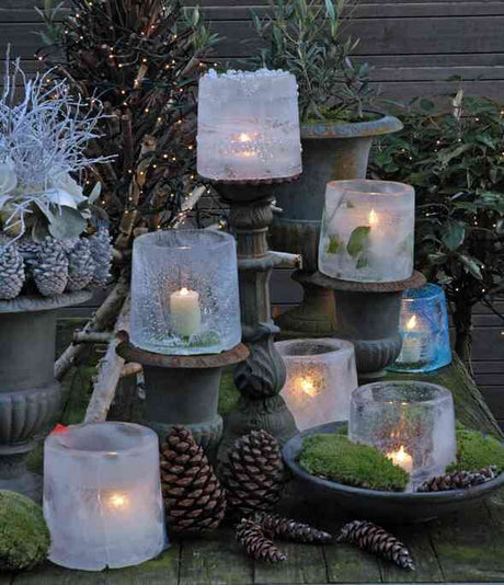 Mehrere Kerzen in Eisgläsern auf alten Pflanzgefäßen, umgeben von Tannenzapfen, Moos und Weihnachtsdekoration.