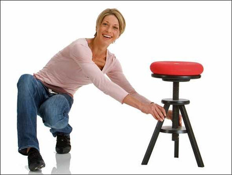 Eine lachende Frau in Jeans und rosa Oberteil sitzt auf dem Boden und hält an einem roten Sitzhocker.