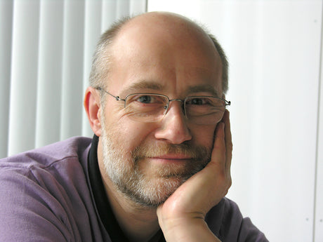 Nahaufnahme eines lachenden Mannes mit Brille, Bart und kurzen Haaren, der die Wange mit der Hand stützt.