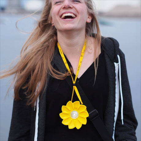 Lächelnde Frau mit gelber Blume als Halskette, trägt schwarze Jacke und hat lange, blonde Haare.