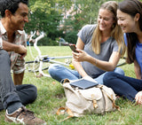 Drei junge Frauen und ein Mann sitzen im Park, lachen und schauen auf ein Smartphone. Fahrrad liegt im Hintergrund.