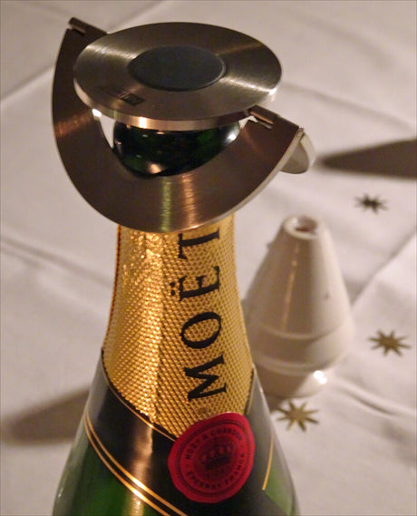 Sektflasche mit goldener Banderole, Metalldeckel, auf einem Tisch mit weißen Tischdecken und goldenen Sternen.