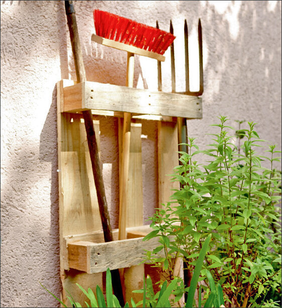 Holzgestell mit Gartengeräten, darunter eine Harke, ein Rechen und eine Schaufel, vor einer Wand mit Pflanzen.