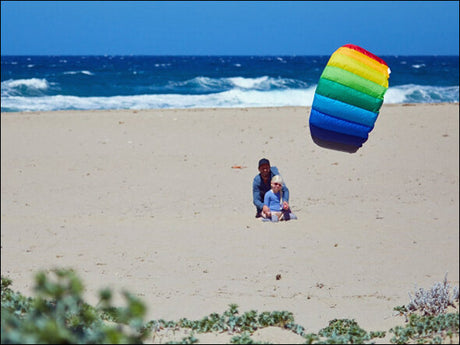 Zwei Personen sitzen im Sand am Strand, neben einem bunten, aufsteigenden Kite, mit Meer und Wellen im Hintergrund.