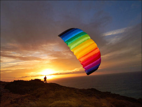 Person mit einem bunten Fallschirm steht auf einem Hügel bei Sonnenuntergang über dem Meer.