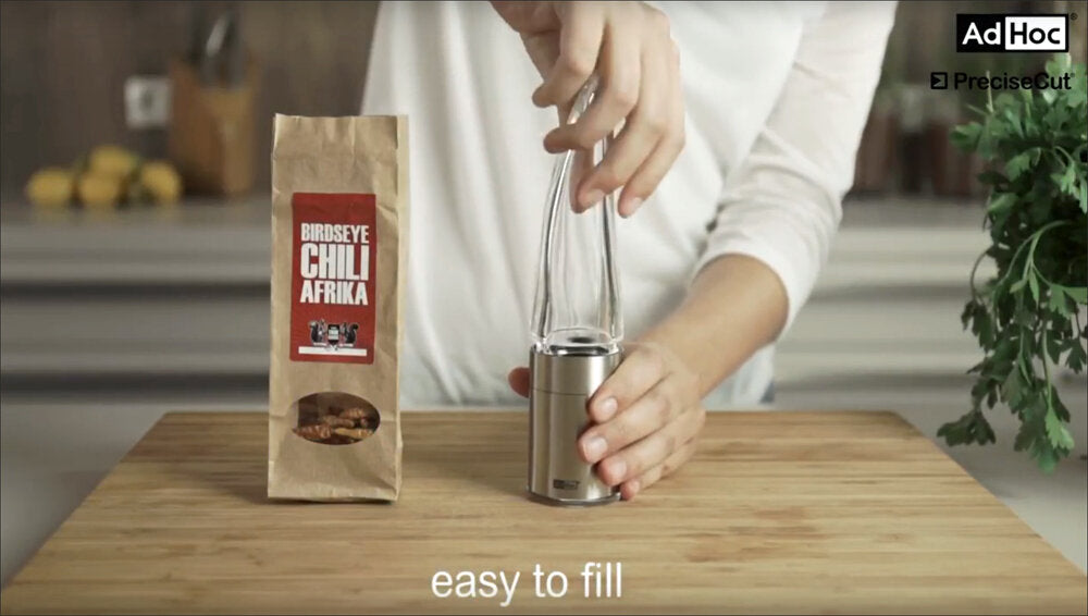 Person mahlt Gewürz aus einer Kaffeemühle, daneben eine Packung "Birdseye Chili Afrika" auf einem Holztisch.