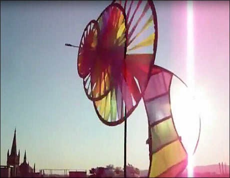 Buntes, bogenförmiges Windspiel mit farbigen Segeln, im Sonnenlicht gegen Himmel und Stadt im Hintergrund.