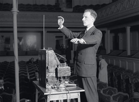 Mann in Anzug dirigiert ein elektronisches Orchester in einem Saal.